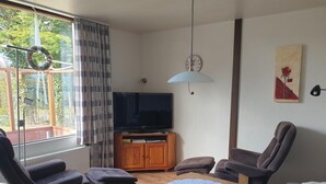 Ett vardagsrum med en tv, trämöbler, ett bord med en spetsduk och ett fönster med gardiner.