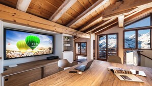 A modern room with a wooden ceiling, large windows with mountain views, a desk with a laptop, and a television displaying a scenic image.