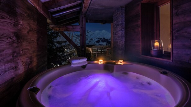 A wooden cabin with a hot tub illuminated by purple lights, a window with a candle, and a view of snow-capped mountains.