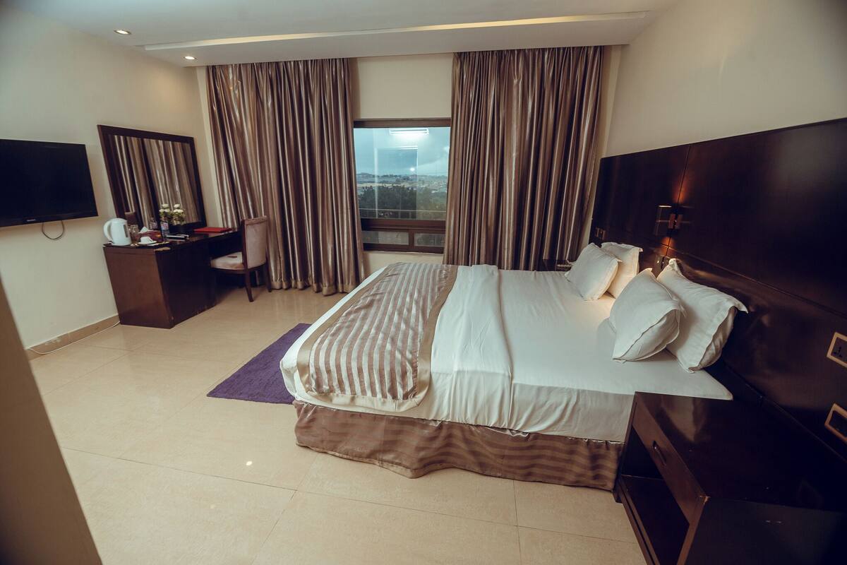 A hotel room with a large bed, a desk with a chair, a television, and a view of the city through the window.