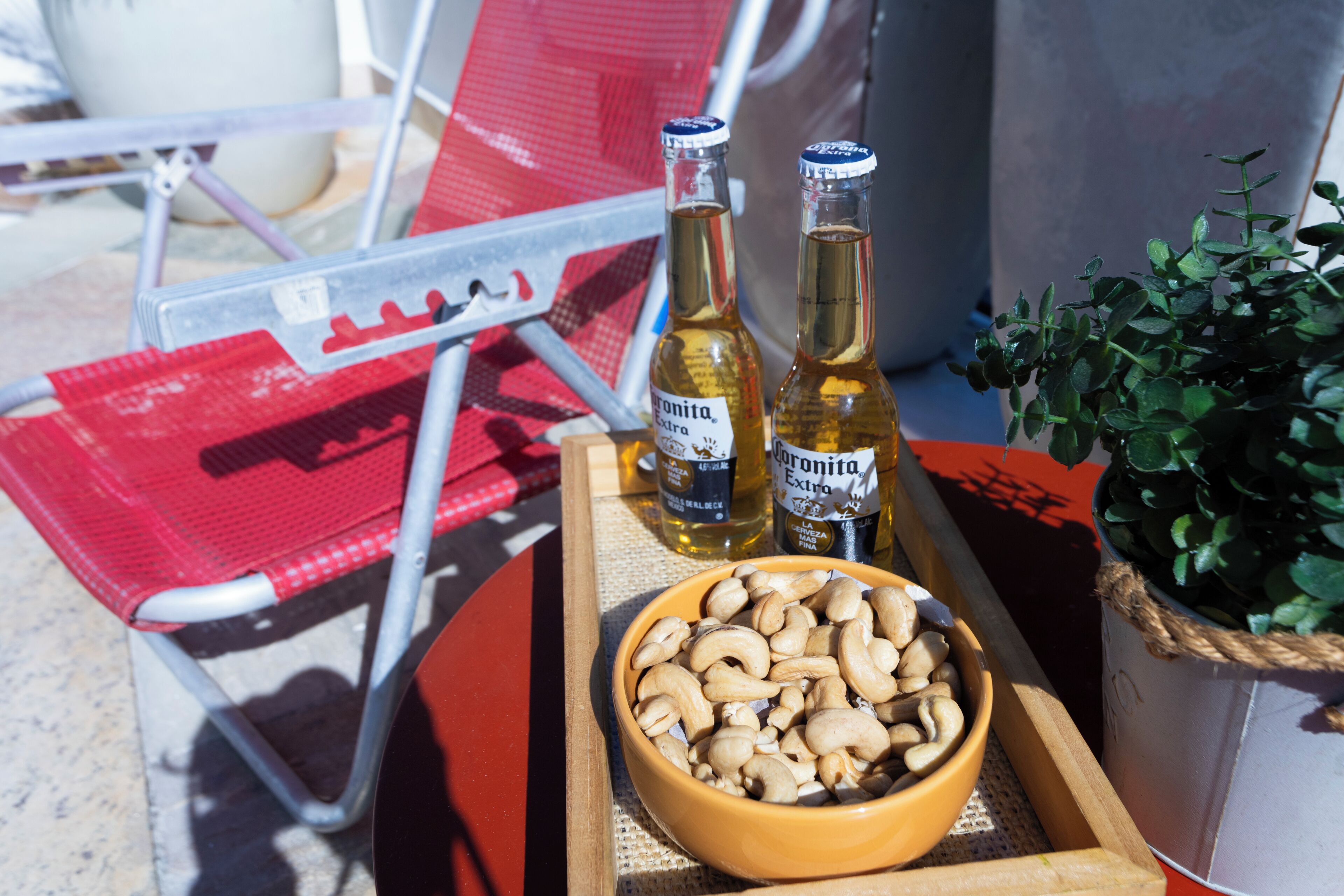 Et fad med to Corona Extra flasker, en skål med cashewnødder, en rød stol og en potteplante.