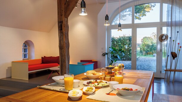 Ein Esstisch ist mit Frühstücksgebäck angerichtet, ein rotes Sofa und ein großes Fenster mit Blick auf Grünflächen.