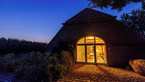 Ein Backsteinhaus mit großem Bogenportal, einem Kopfsteinpflasterweg, der davor führt, und einem Garten mit Pflanzen und einem Baum.