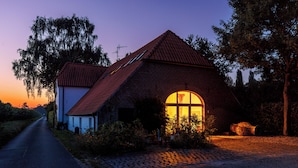 Ein Haus aus Backstein mit rotem Dach und einem großen Bogenfenster, umgeben von Bäumen und einem Kopfsteinpflasterweg.