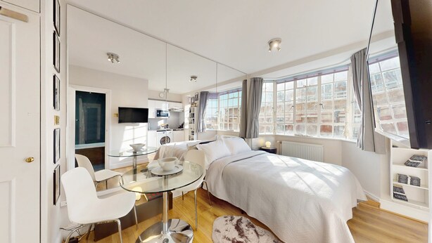 A modern hotel room with a bed, a glass table, a chair, a TV, and a view of the city through large windows.