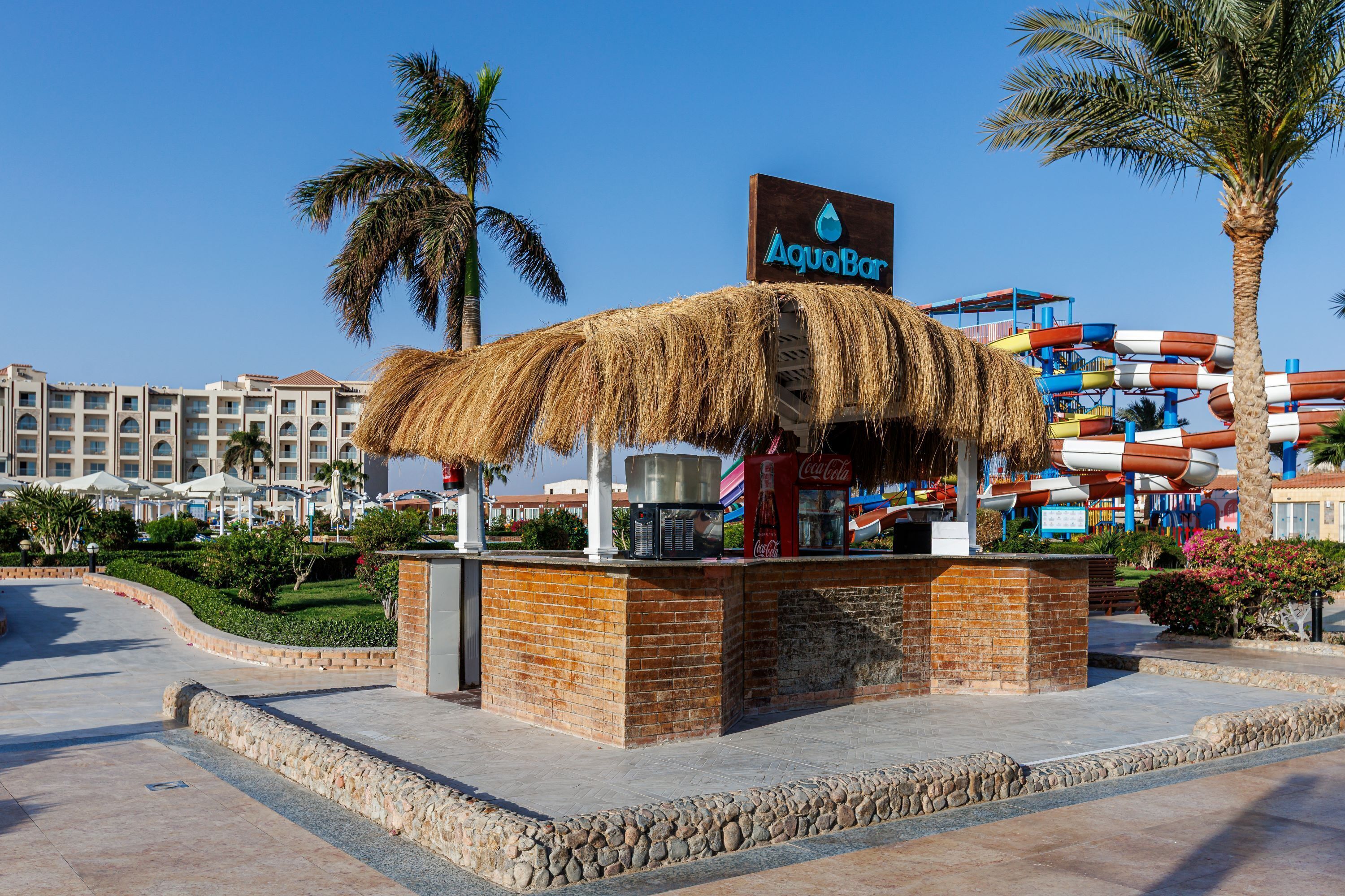 Un bar à toit de chaume appelé AquaBar, avec une enseigne Coca-Cola, situé devant un parc aquatique et des bâtiments d'hôtel.