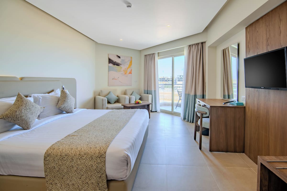 A hotel room with a large bed, a desk, a chair, a TV, and a balcony with curtains.