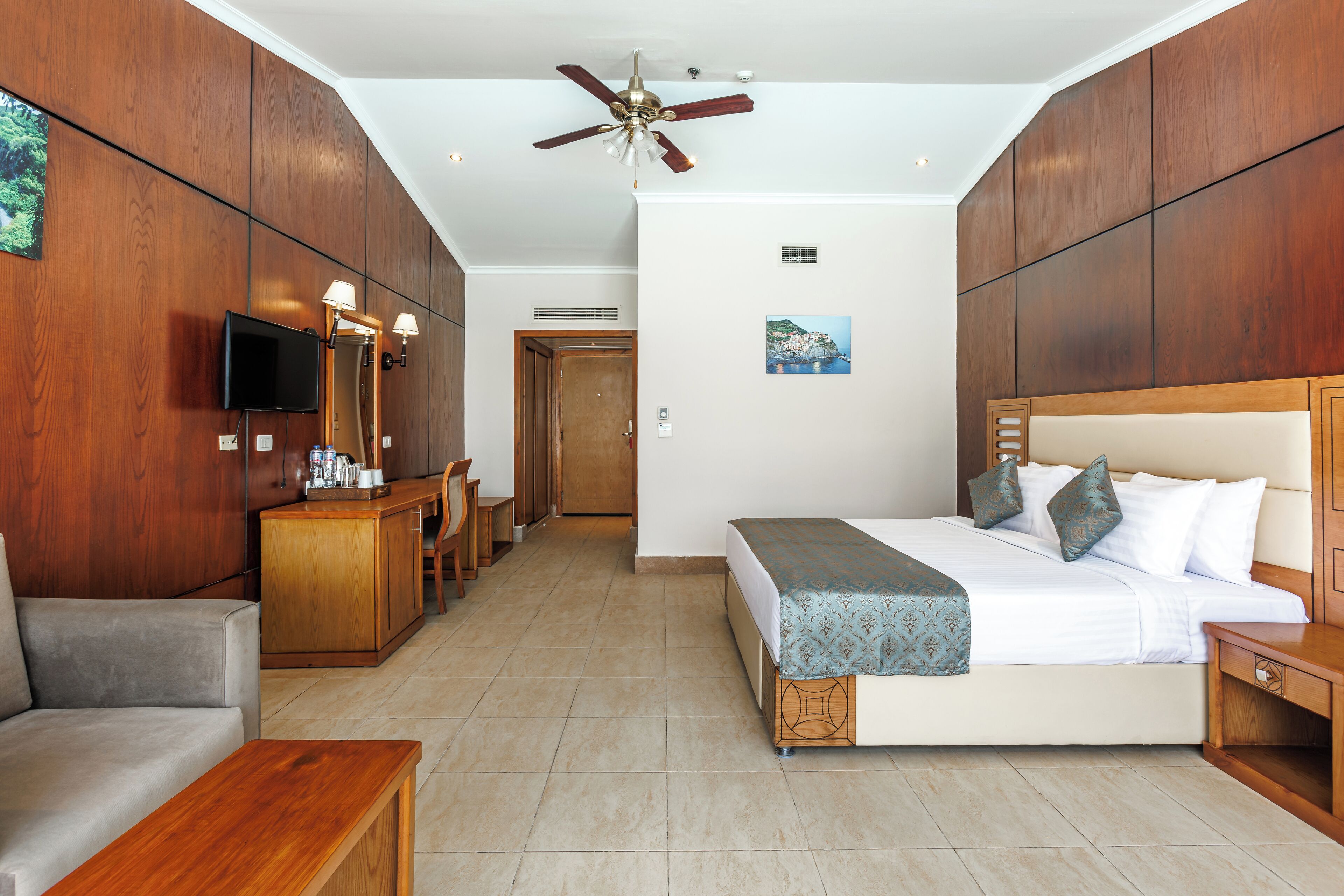 A hotel room with a bed, a desk, a chair, a sofa, a television, and a ceiling fan.
