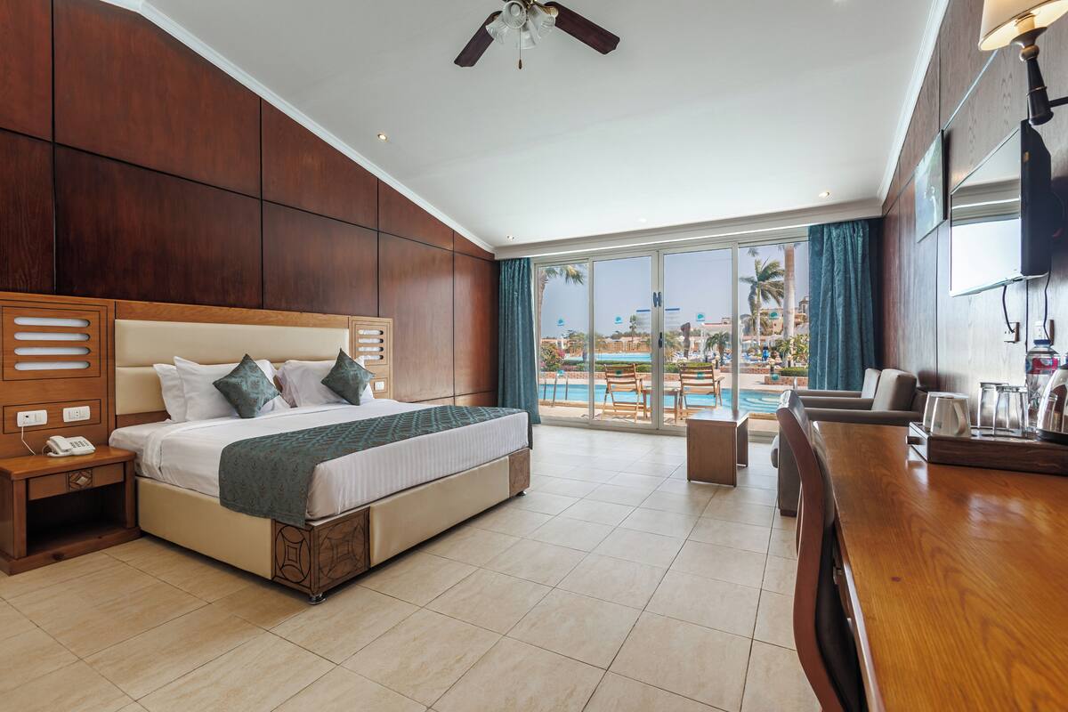 A modern hotel room with a large bed, a balcony with a pool view, and a wooden bar area.
