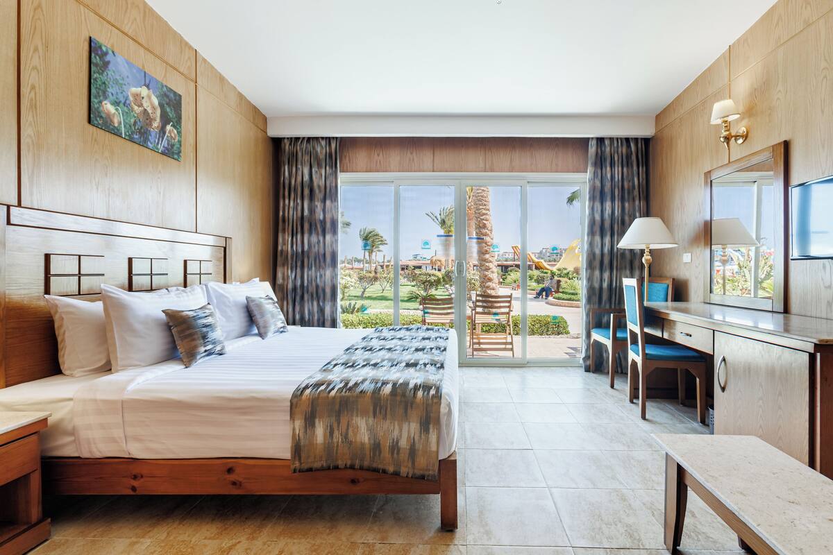A hotel room with a large bed, a desk, a chair, and a view of a resort area.