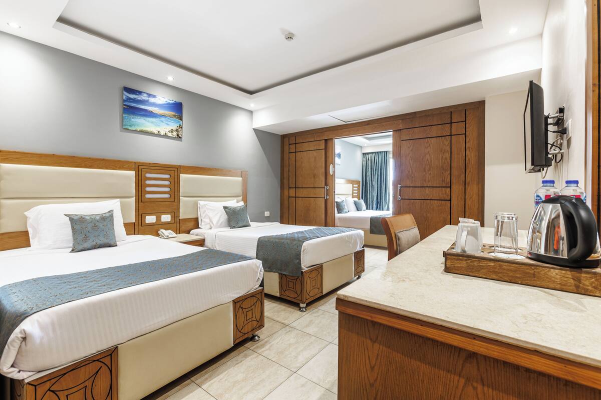 A hotel room with two beds, a wooden headboard, a desk with a kettle, and a TV.