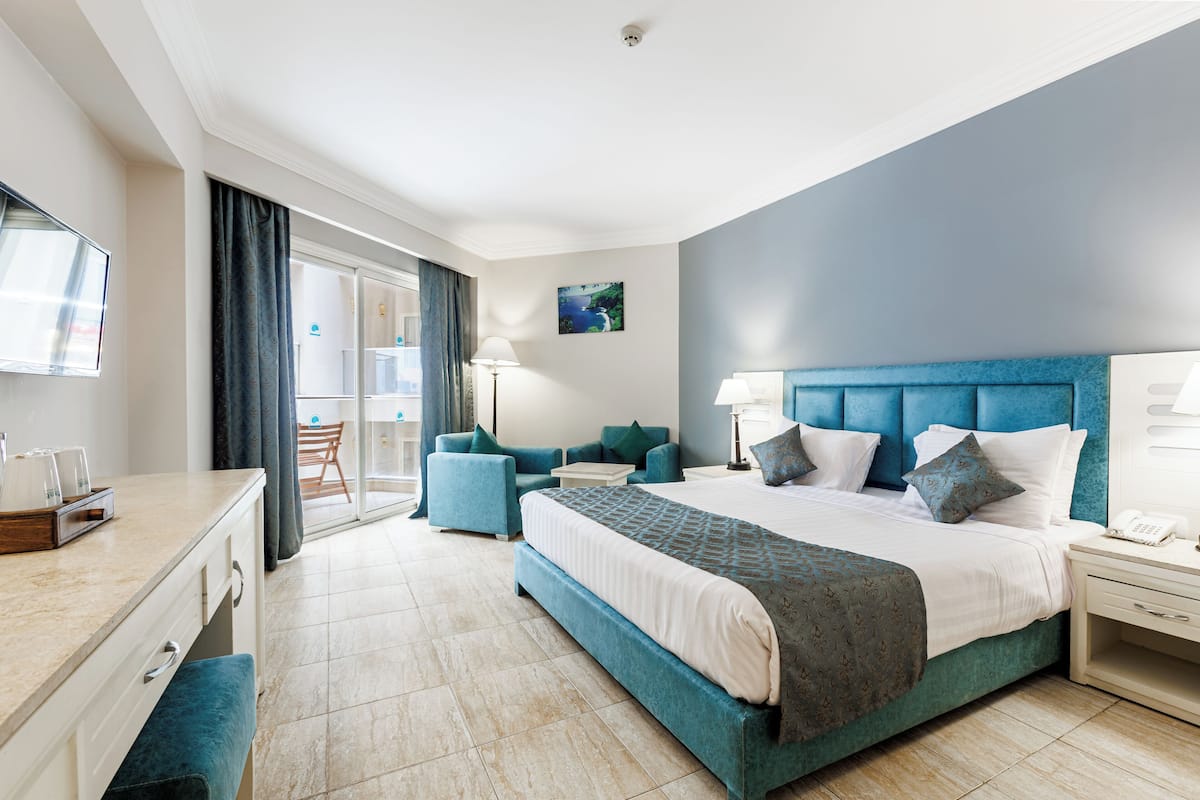 A modern hotel room with a large bed, two armchairs, a desk, and a view of the outdoors.