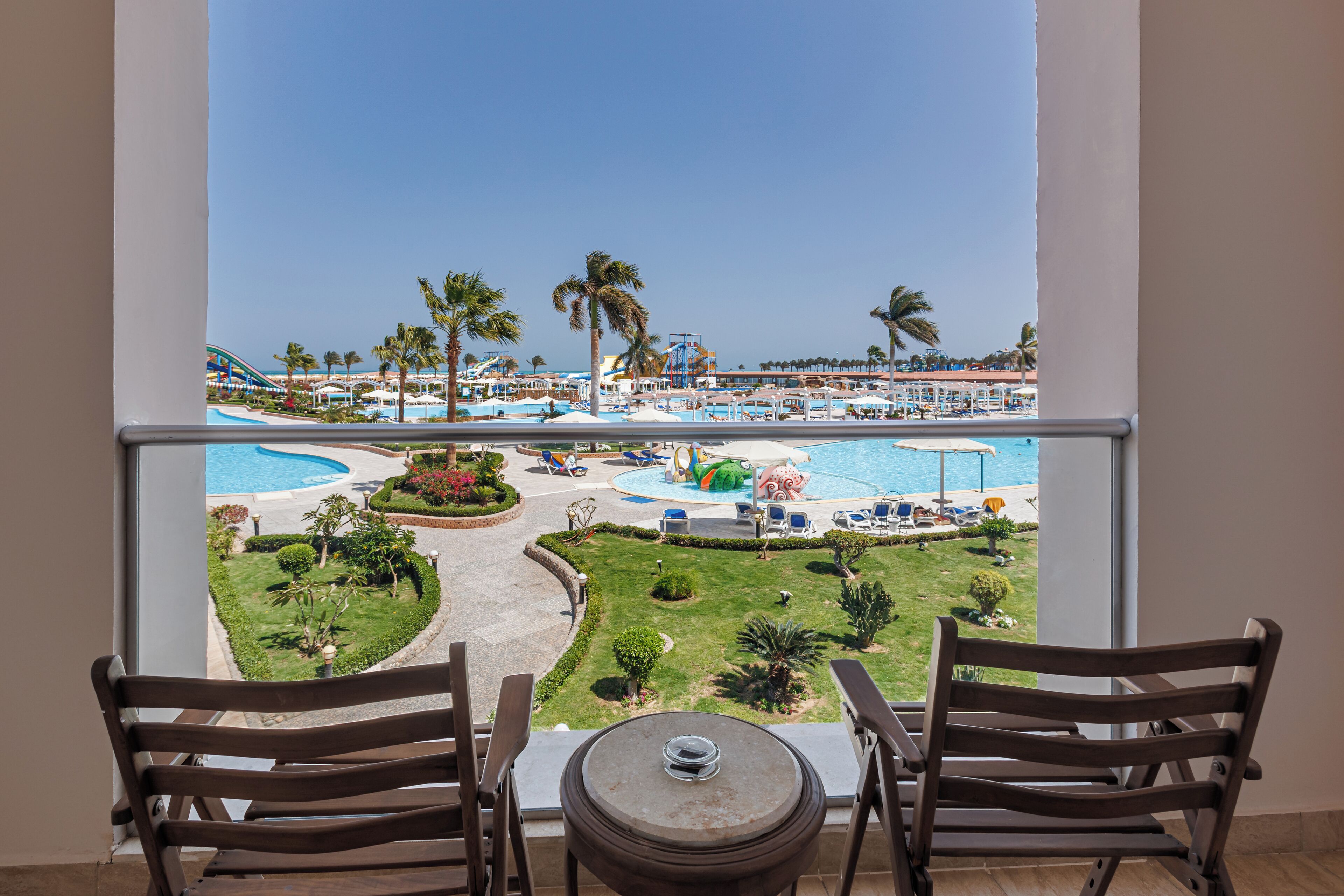A balcony with two wooden chairs and a small table, overlooking a pool area with palm trees and a water slide.