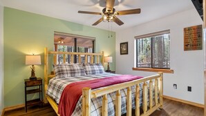 Un dormitorio con una cama de madera, dos mesitas de noche con lámparas, un ventilador de techo y ventanas amplias que ofrecen vistas al exterior.