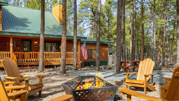 Una cabaña con fogón rodeado de sillas Adirondack, una terraza y vistas a altos pinos.