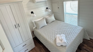 Un dormitorio con techo de madera, paredes blancas, cama con colcha acolchada, dos lámparas de noche, unidad de aire acondicionado y ventana con persianas.