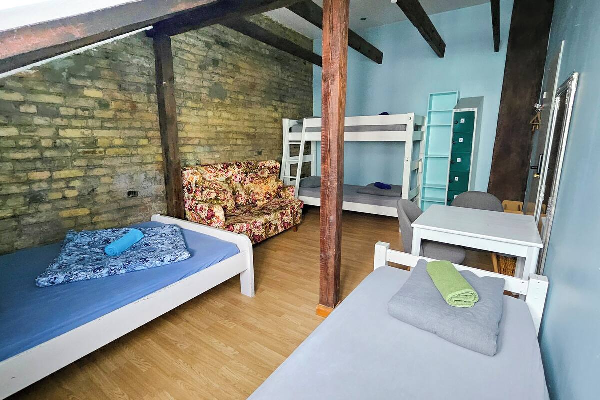 A dormitory room with bunk beds, a wooden floor, a brick wall, a floral sofa, and a desk.