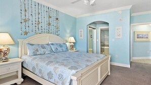 Un dormitorio con cama, mesitas de noche, ventilador de techo y una puerta que da a otra habitación.