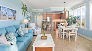 Un salón espacioso con un sofá azul, sillones blancos, una mesa de comedor con sillas blancas, una cocina al fondo y un ventanal con vistas.