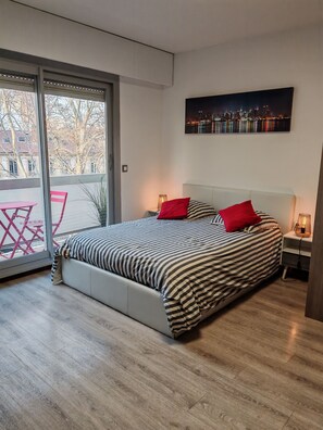 Une chambre moderne avec un grand lit, un tableau représentant un paysage urbain et une porte-fenêtre coulissante donnant sur un espace extérieur.