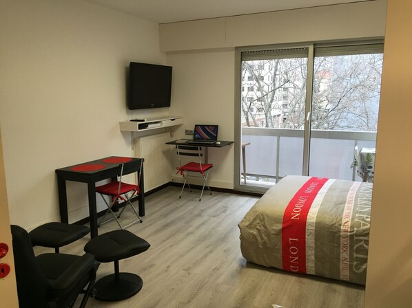 Une chambre d’hôtel avec un lit, un bureau équipé d’un ordinateur, une télévision et un balcon donnant sur la verdure.