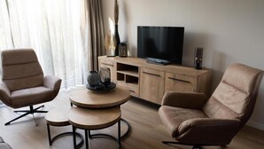Un salon moderne avec deux fauteuils beiges, une table basse en bois et un téléviseur à écran plat posé sur un meuble en bois.