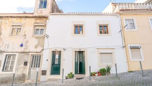 Une rangée de bâtiments aux styles architecturaux variés, dont un immeuble blanc avec une porte et des fenêtres vertes, ainsi que des bâtiments plus anciens adjacents, dont on peut voir les signes d’usure.