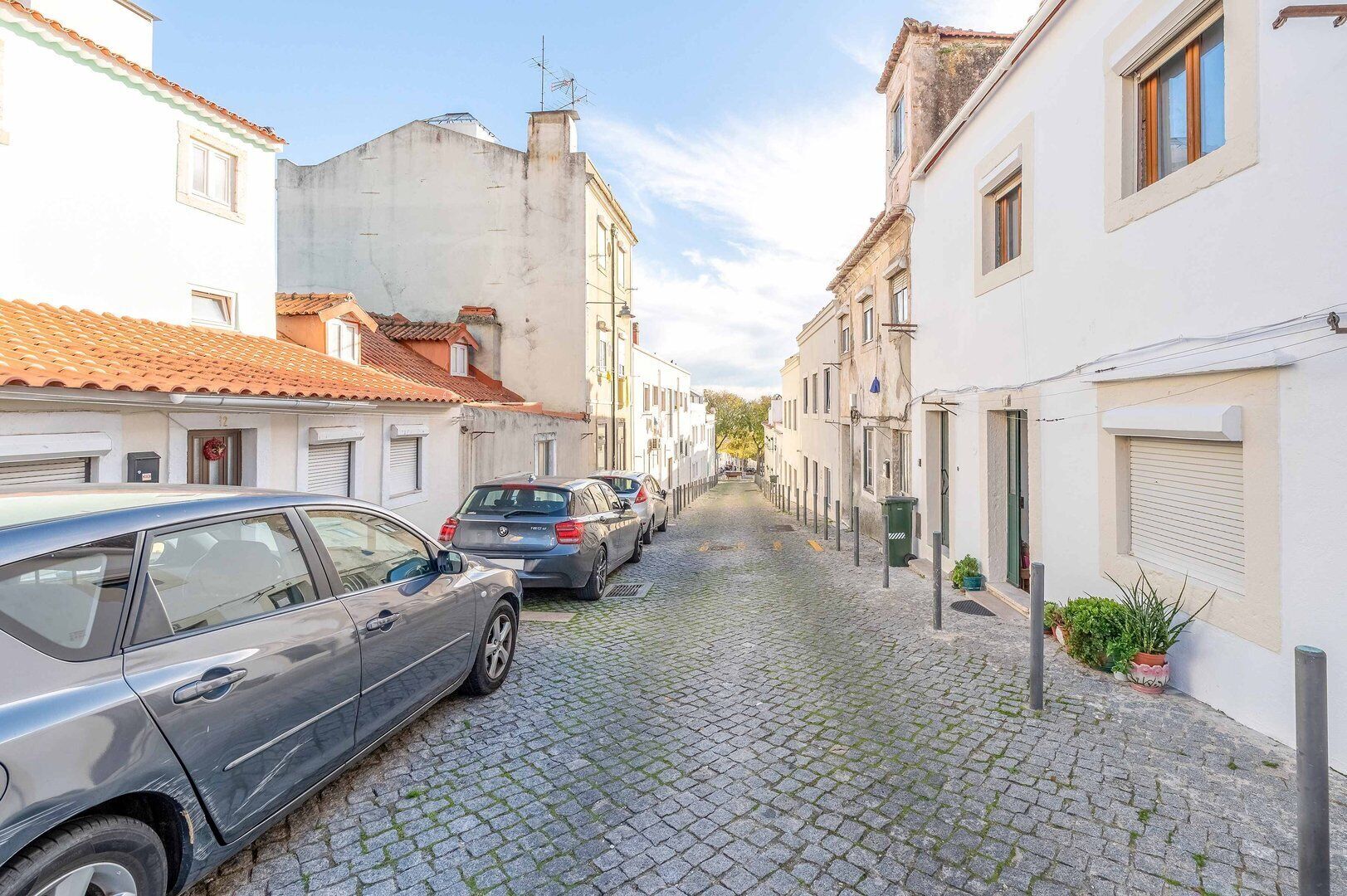 Una estrecha calle adoquinada flanqueada por edificios blancos, coches aparcados y un cielo despejado.