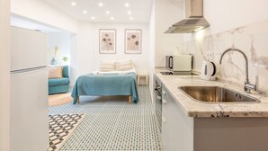 Une chambre d’hôtel compacte avec une kitchenette, un lit recouvert d’une courtepointe bleue, un sol à motifs et deux œuvres d’art encadrées accrochées au mur.