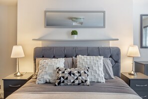 Ein ordentlich eingerichtetes Schlafzimmer mit einem grauen, bezogenen Kopfteil, zwei Nachttischen mit Lampen und einer kleinen Topfpflanze auf einem Regal über dem Bett.