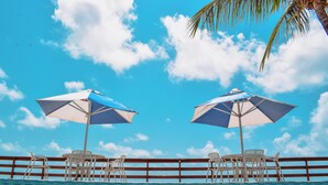 Zona de piscina con dos sombrillas, sillas blancas y barandilla de madera.
