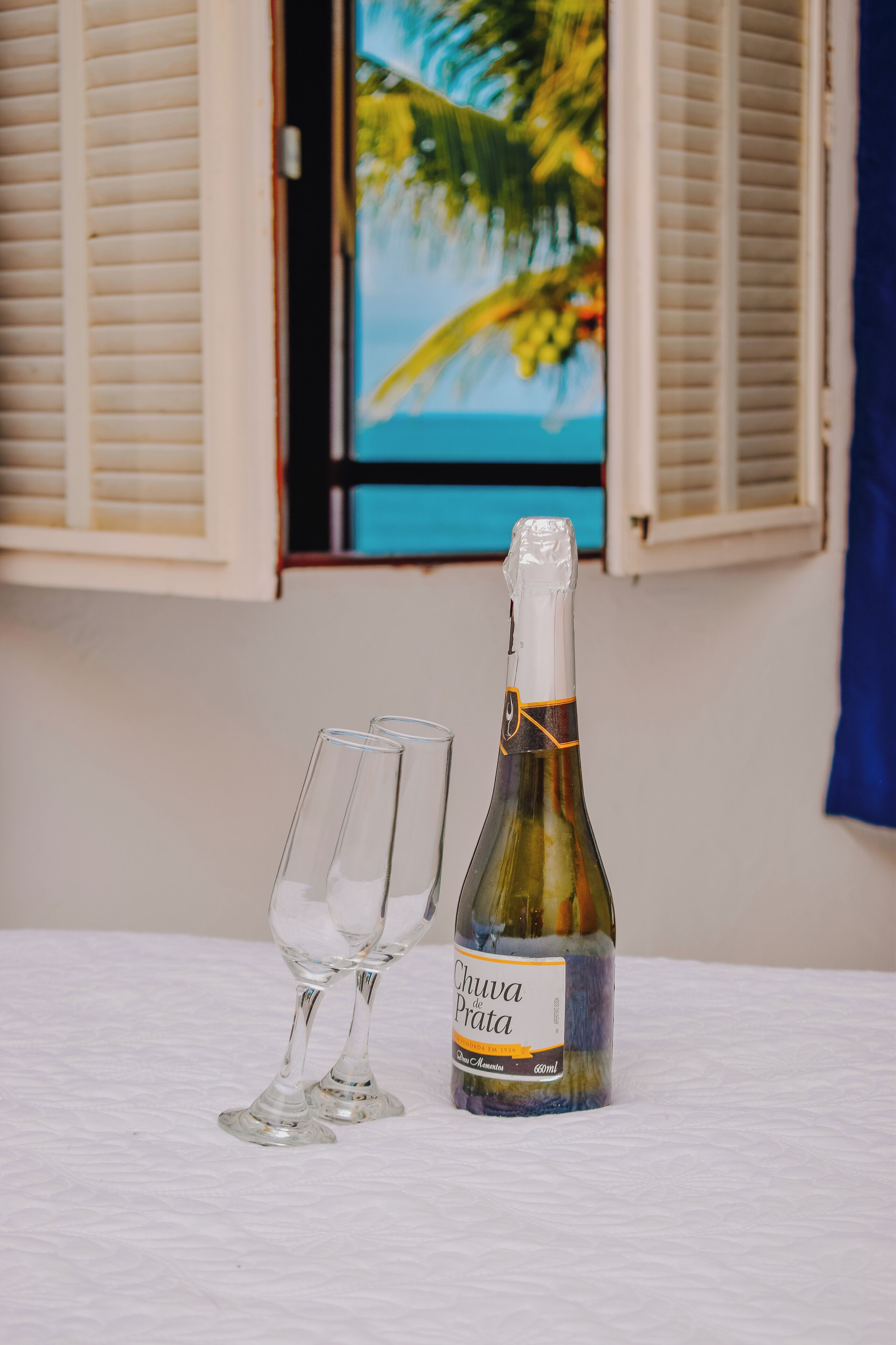 A bottle of Chuva de Prata champagne and two glasses on a table with a view of a window with shutters and a tropical painting.