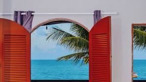 Una habitación de hotel con una cama, una ventana con contraventanas rojas y vista al océano.
