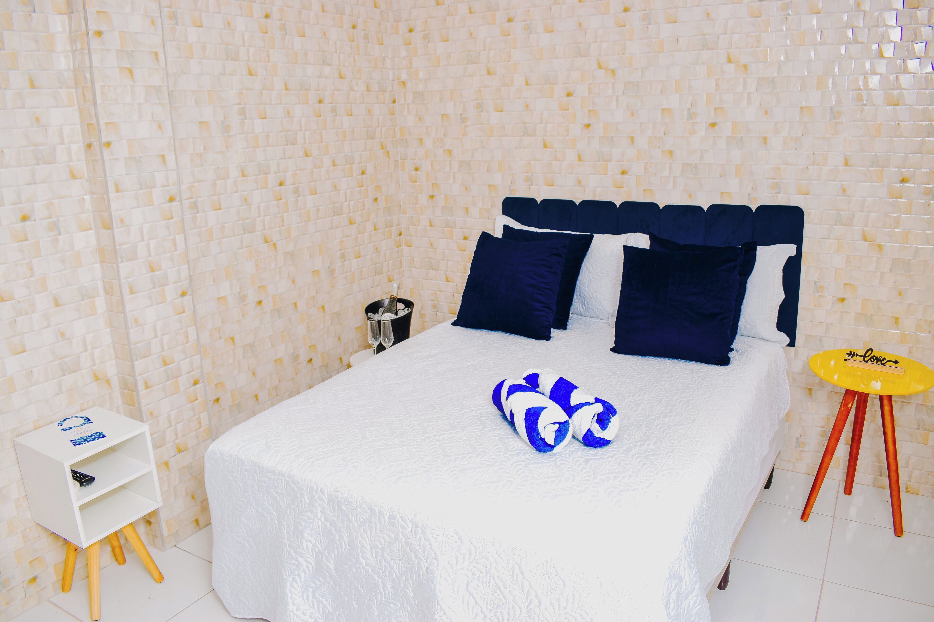 A neatly made bed with a white bedspread and blue and white pillows, a small white nightstand with a remote control, a pair of blue and white towels, and a yellow stool with a logo.