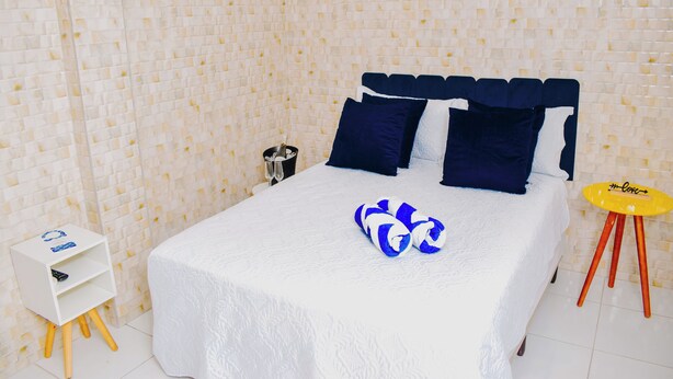 Una cama bien tendida con una colcha blanca y almohadas azules y blancas, una mesita de noche pequeña de color blanco con un control remoto, un par de toallas azules y blancas, y un taburete amarillo con un logotipo.