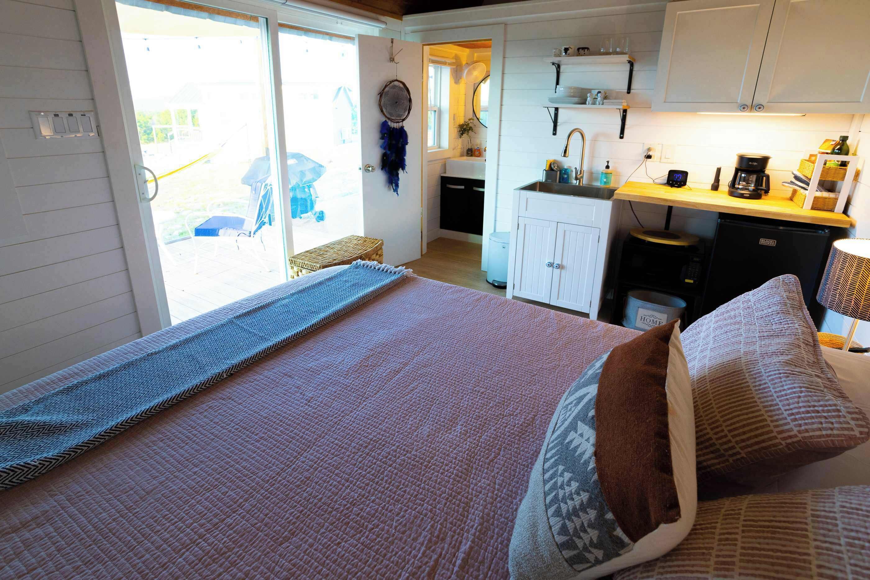 A bedroom with a bed, pillows, and a view of a kitchen and balcony.