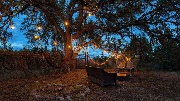 Una hamaca bajo un árbol con luces de cuerda, dos sillas de madera y un entorno rústico.