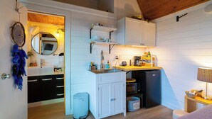 Cocina compacta con armarios blancos, un fregadero, estufa y refrigerador. Hay un pequeño cubo de basura y una estantería con utensilios de cocina.