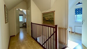A hallway with wooden flooring, a wooden railing, a painting on the wall, and a window with curtains.