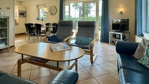 A modern living room with a sofa, a coffee table, and a TV. There is a dining area with chairs and a table.