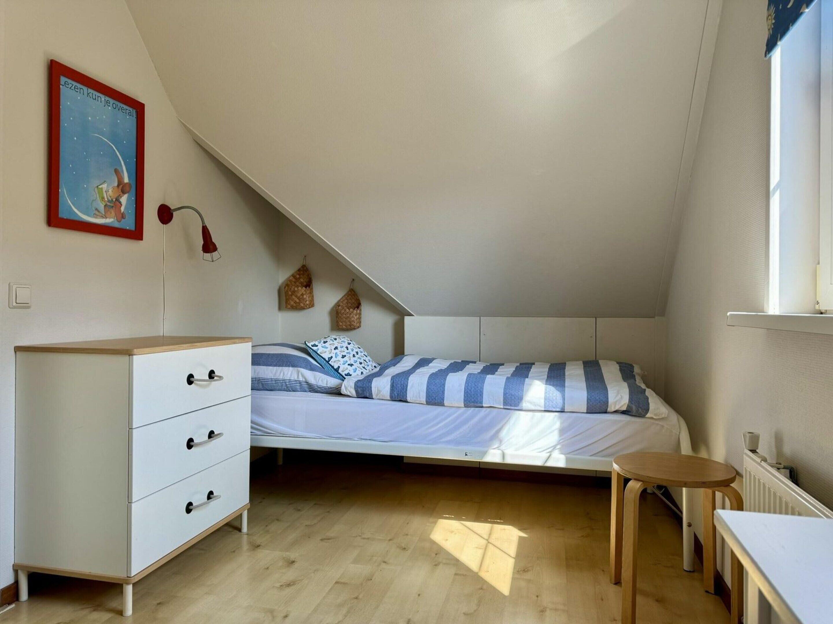 A small, well-lit attic bedroom with a bunk bed, a white dresser, a small table, and a radiator.