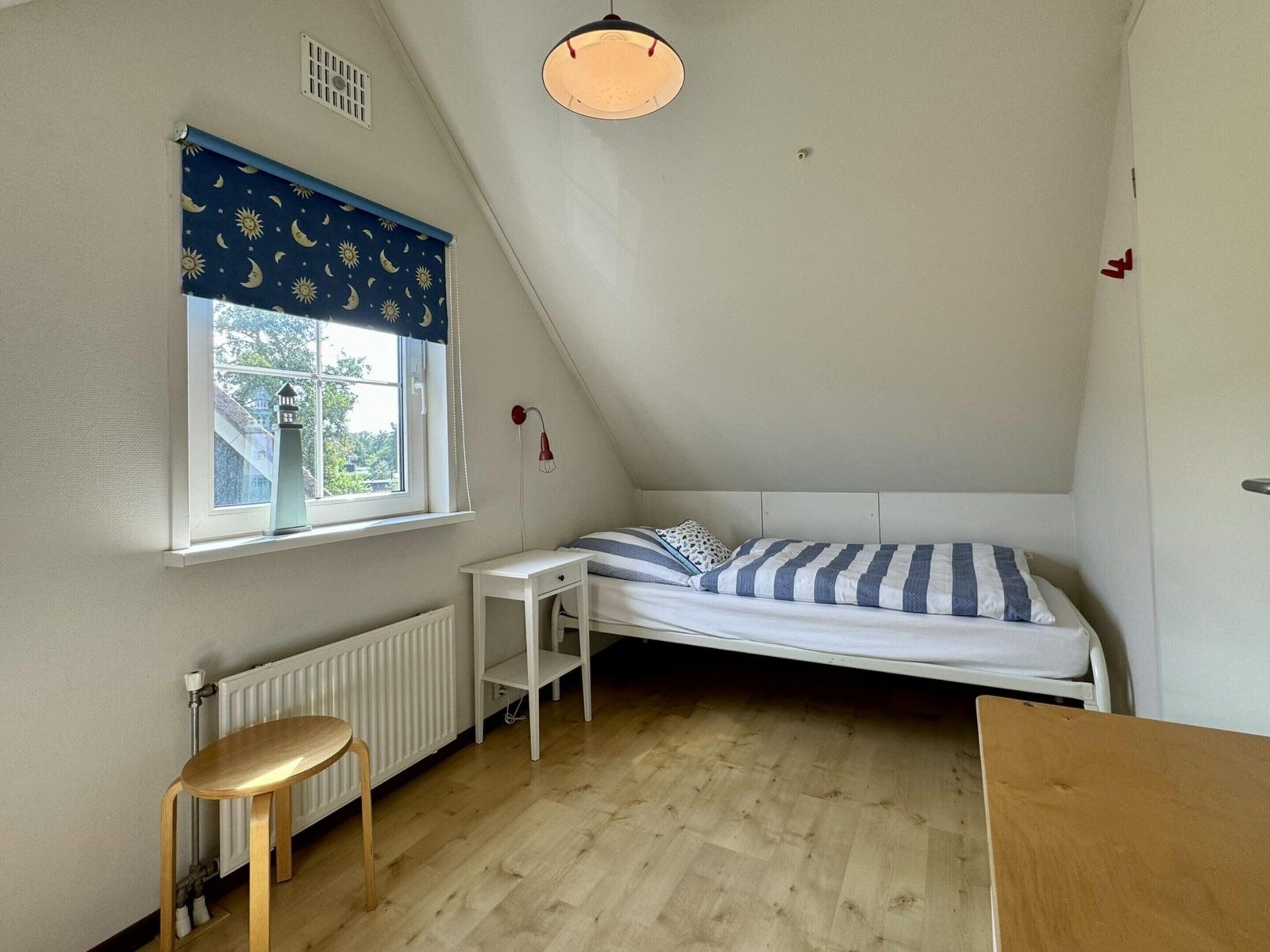 Une petite pièce triangulaire avec un plafond en pente, un lit simple avec une literie à rayures, une petite table, un tabouret, un radiateur et une fenêtre ornée d’un motif représentant la lune et le soleil.