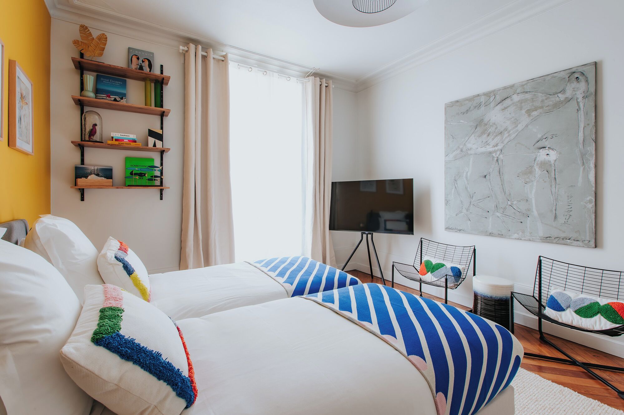 Et moderne hotelværelse med en seng, et fjernsyn, et stort abstrakt maleri og en reol med bøger og pynt.