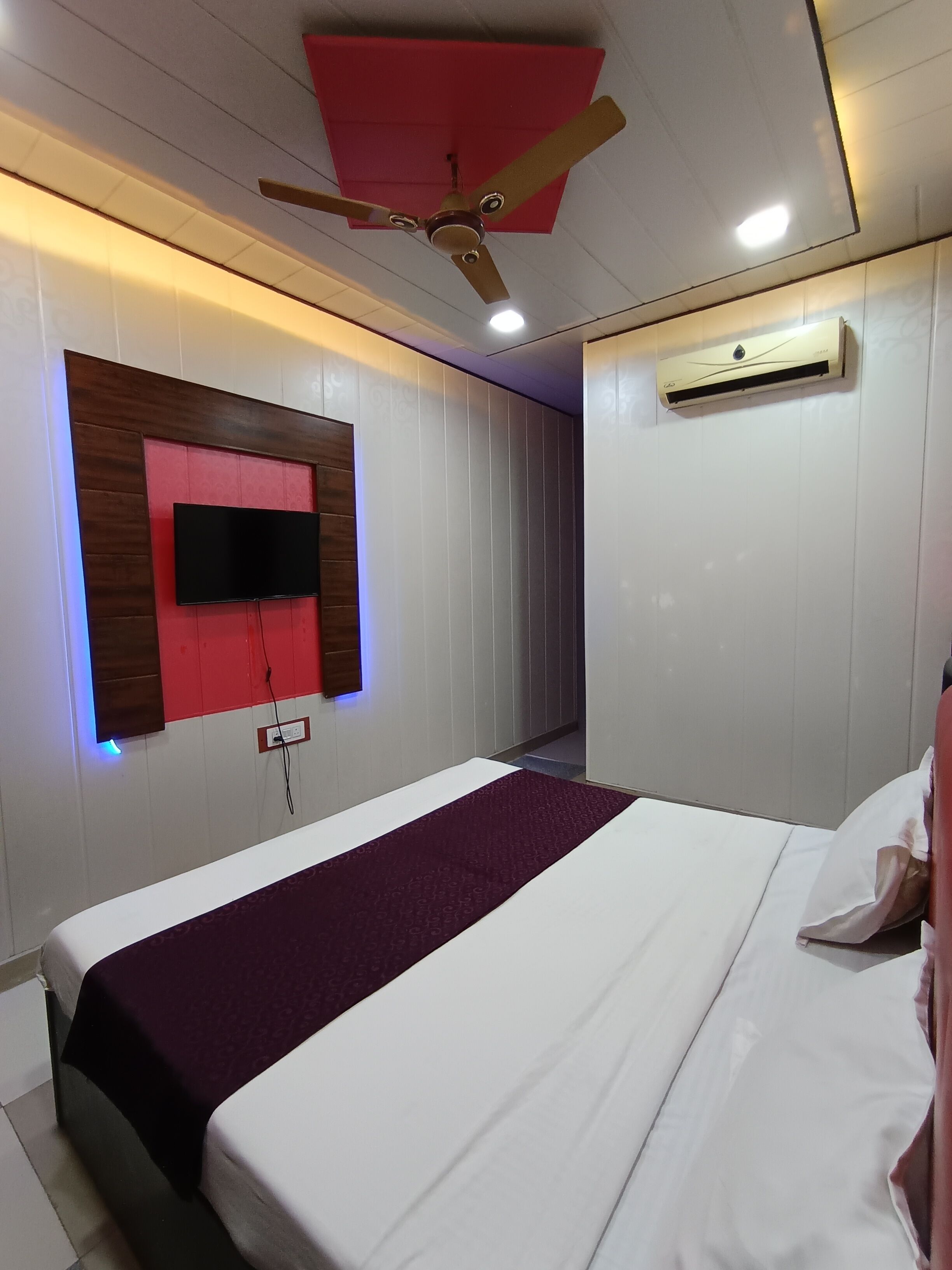A modern hotel room with a bed, a mounted TV, a ceiling fan, and an air conditioning unit.