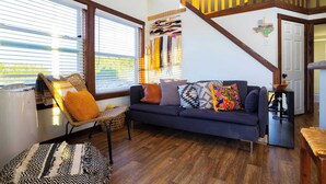Uma sala de estar aconchegante com um sofá azul escuro, uma poltrona de vime e um tapete estampado. Uma escada com corrimão de madeira leva a um andar superior.
