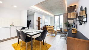 A modern kitchen with white cabinets, a dining area with a black table and chairs, and a city view through large windows. - Hoola Cove Executive One Bed Apartment (London)