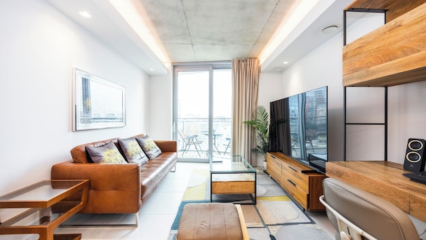 A modern living room with a sofa, coffee table, ottoman, TV, and wooden sideboard. - Hoola Cove Executive One Bed Apartment (London)
