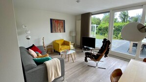 A modern living room with a grey sofa, a yellow armchair, a unique black chair, a flat-screen TV, and a large window offering a view of the outdoors.