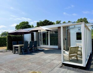 A covered patio area with a table and chairs, a glass enclosure, and a brick wall. - Lovely Bungalow With Huge Garden (Sint Maartensvlotbrug)