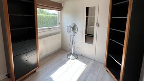 A room with a built-in wardrobe, a standing fan, and a window with a view of greenery.